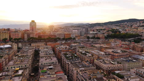drone lateral shot over genoa rooftops at sunset, showing old colorful buildings, a tall modern block in the distance, and mountains behind the city