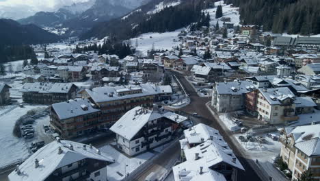 Vista-Aérea-De-Drones-Volando-Sobre-El-Paisaje-Urbano-Invernal-En-Val-Di-Fassa,-Italia,-Con-Edificios-Cubiertos-De-Nieve-Y-Dolomitas-Como-Telón-De-Fondo