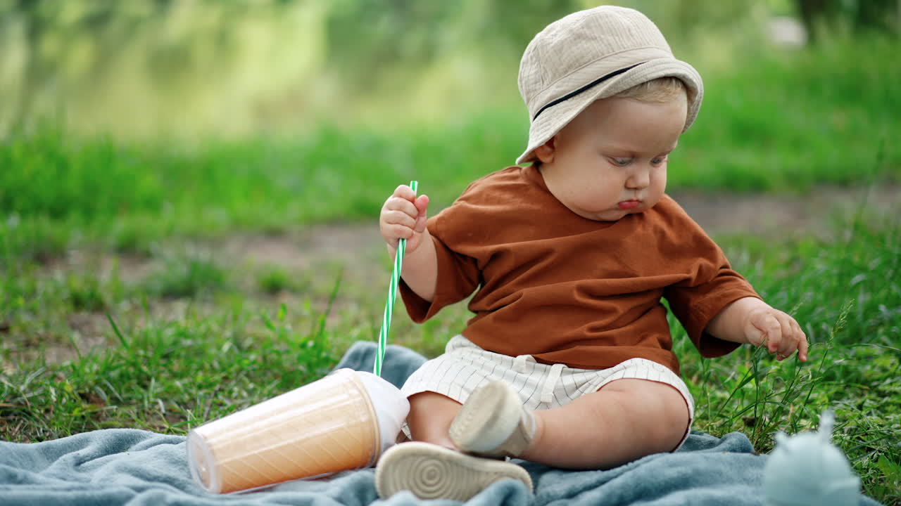 Cute little toddler boy wearing a brown t-shirt, white shorts and a cap sitting on the plaid. Kid holds a straw from a cup and plays with grass.