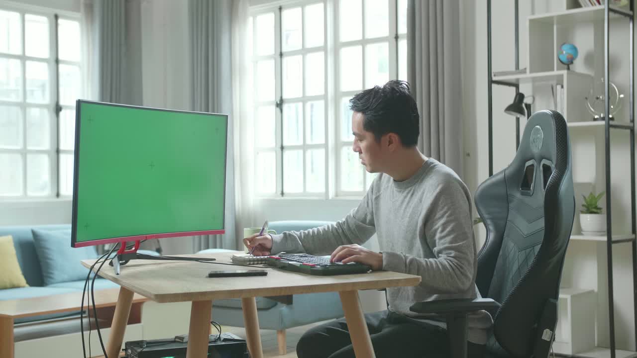Asian Man In Long Sleeved T-Shirt And Black Pants Looking At Green Screen Desktop Computer And Writing In Notebook On A Table While Working At Home.