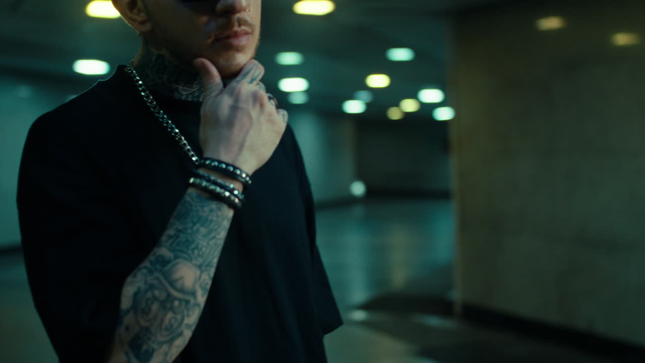 A man with tattoos and sunglasses posing in a dimly lit urban underpass