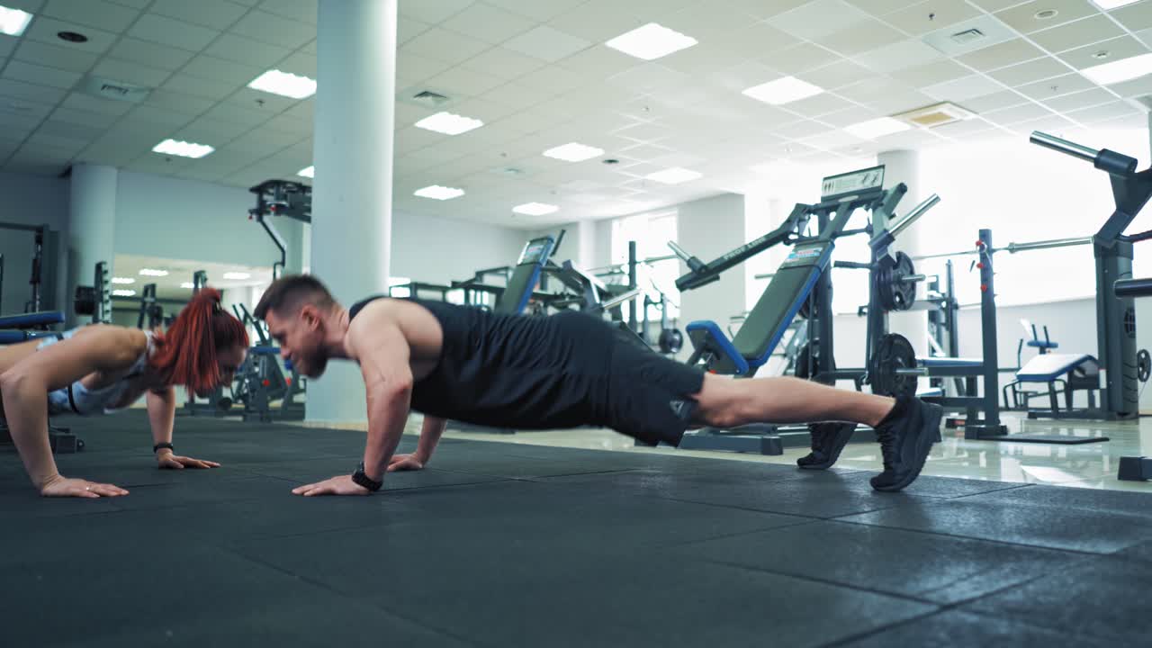 Fit couple doing push ups together in gym. Young sporty man and woman doing push ups and clap each other on the palm. Fitness studio.