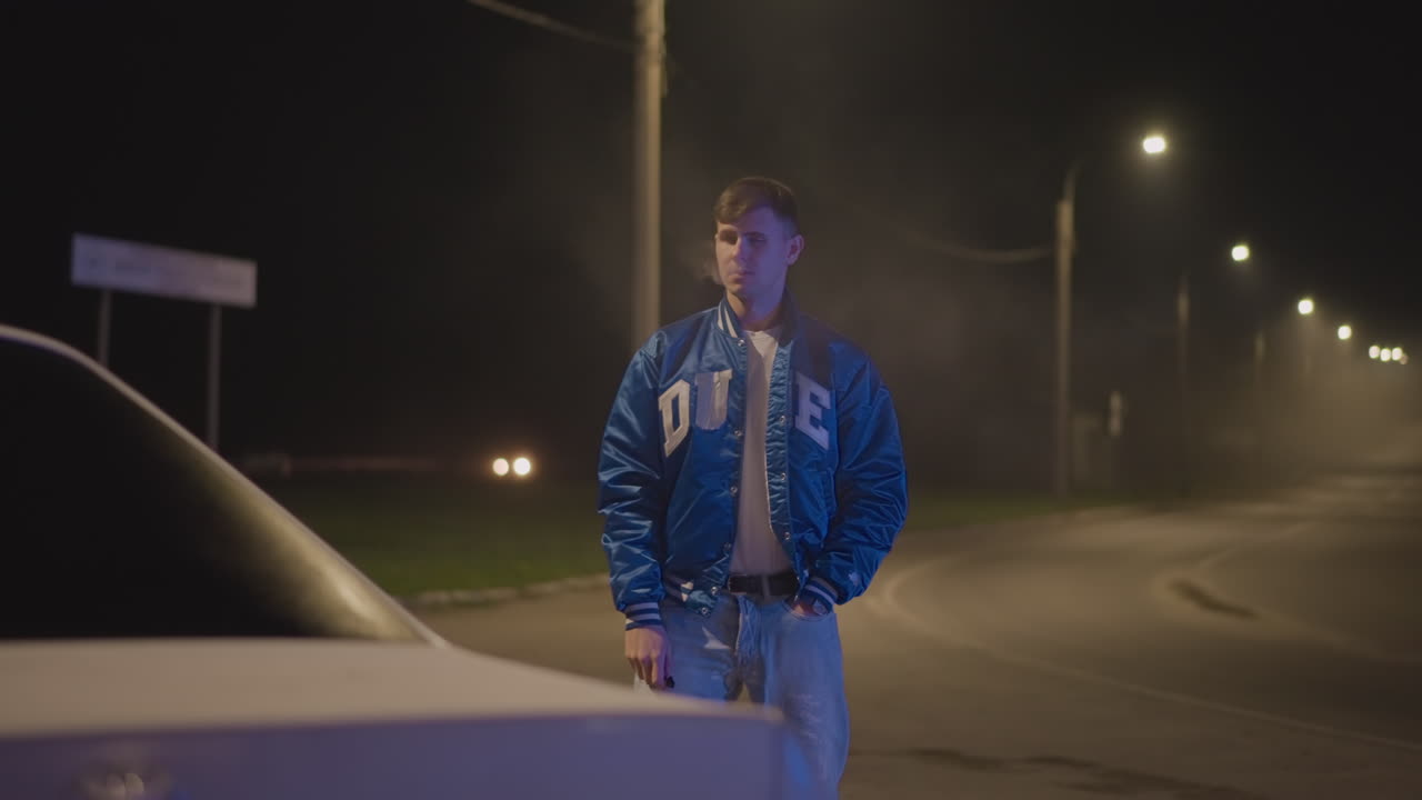 nattlig veikantscene, hvit mannlig sjåfør i collegejakke lener seg mot panseret på bilen og puster røyk ut i den kalde luften under en rekke gatelys på en tom, svingete vei, stemningsfull, filmatisk atmosfære av ensomhet, tåke