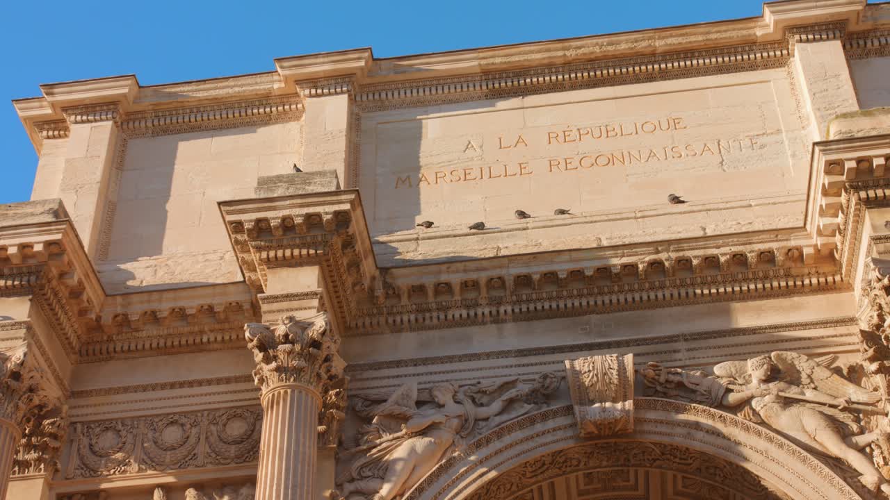 Vista dell'arco di trionfo a Marsiglia, che cattura dettagli architettonici e iscrizioni sotto cieli azzurri e sereni. Le riprese diurne evidenziano il significato storico del luogo.