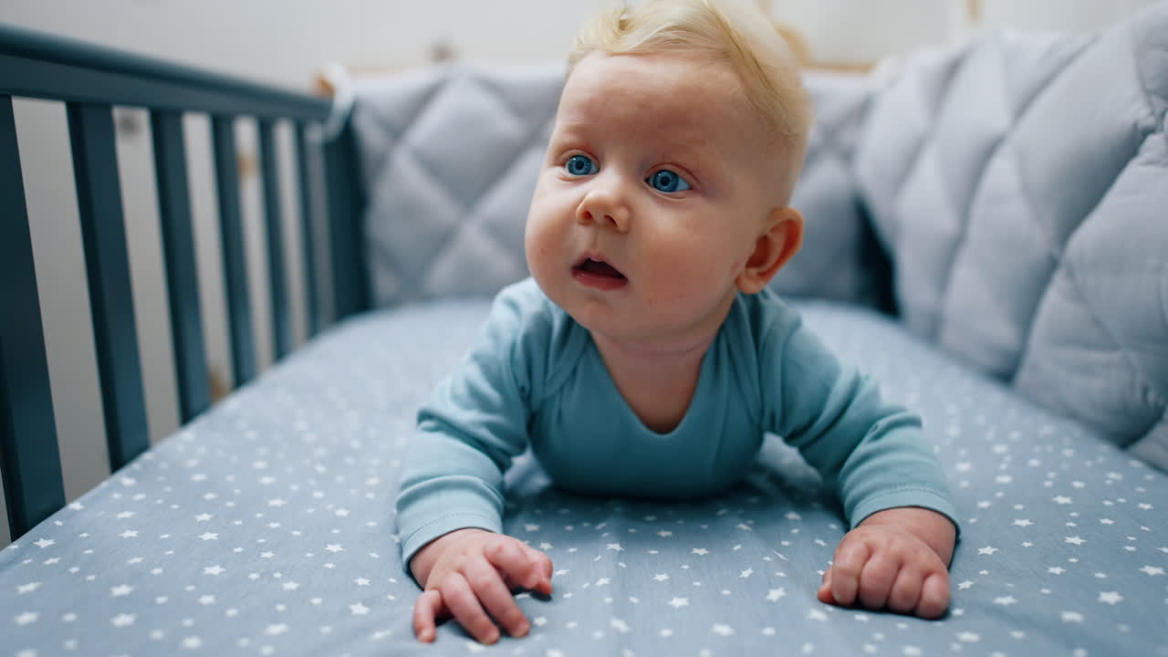 Blonde baby boy in blue clothes lies on belly leaning on his hands. Adorable kid looks up with surprise.