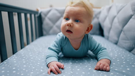 blonde baby boy in blue clothes lies on belly leaning on his hands. adorable kid looks up with surprise.