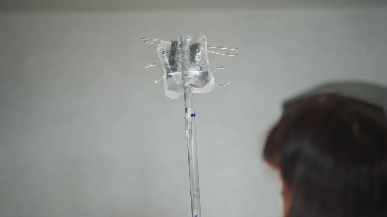 窓際で点滴スタンドの近くで休む人。穏やかな光の雰囲気の中で点滴器具の隣に座る患者。柔らかく照らされた部屋で、点滴スタンドの近くに穏やかに座る白人患者。
