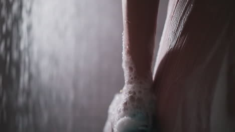 woman washes body side with loofah in shower cabin closeup. lady cleans body skin with foamy mesh sponge in water sprays. pampering and hygiene