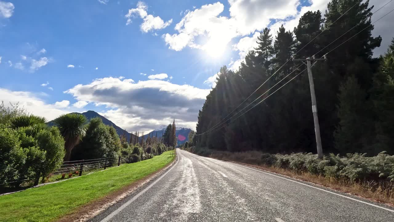 Vehicle drives on sunlit rural road, passing trees and mountains under partly cloudy sky