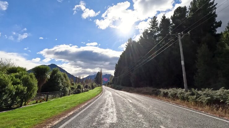 Veicolo percorre una strada rurale soleggiata, superando alberi e montagne sotto un cielo parzialmente nuvoloso