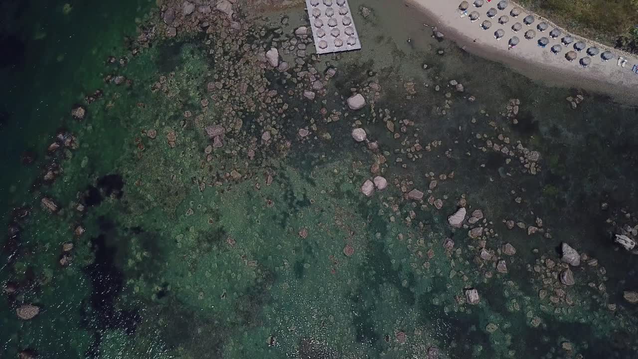 Drone view over seawaves revealing rocky beach and straw umbrellas near bar