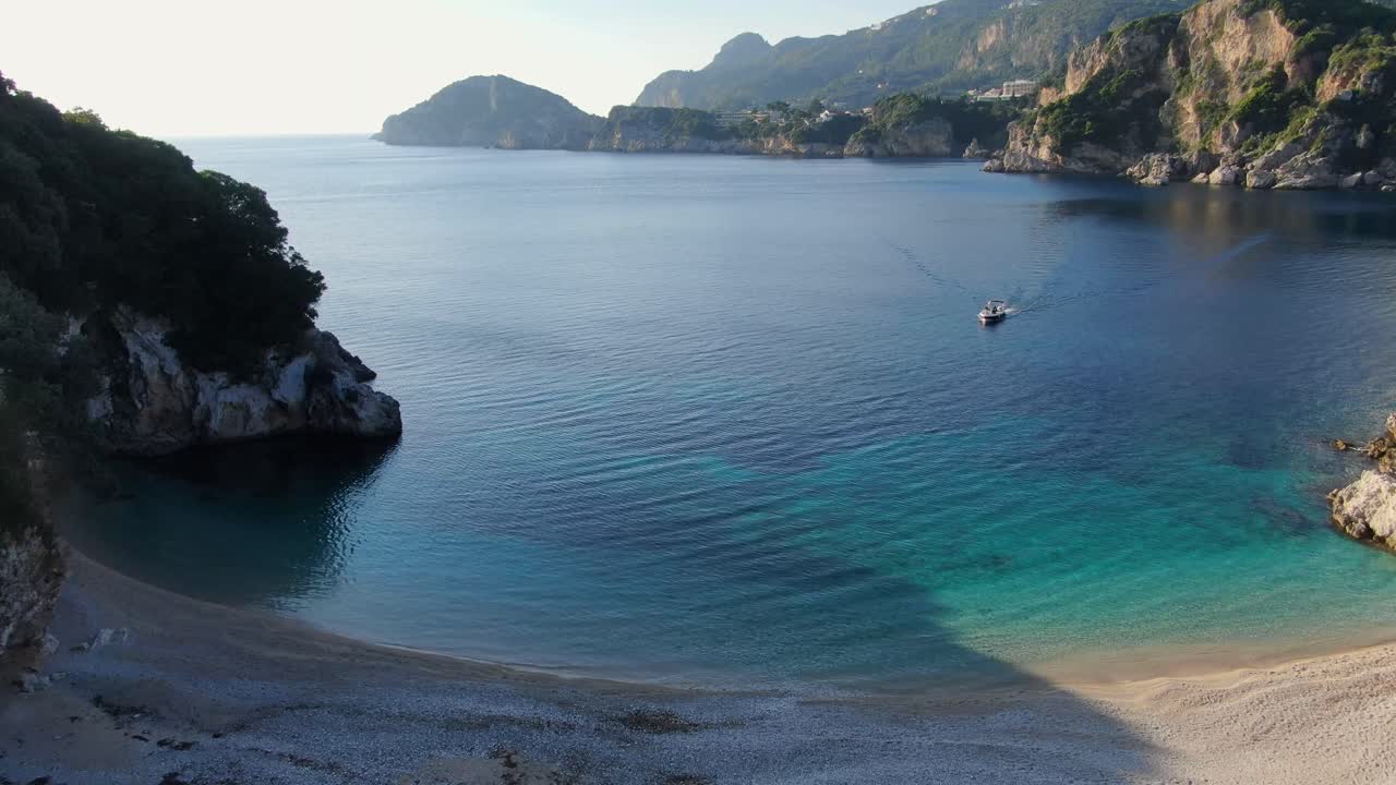 Aerial drone view flying over a stunning secluded beach cove in Paleokastritsa, Corfu, Greece, with turquoise water, cliffs, and a small boat