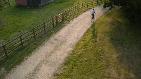 Toma-Aérea-De-Un-Dron-De-Una-Mujer-Montando-En-Bicicleta-Por-Un-Camino-Rural-Al-Atardecer