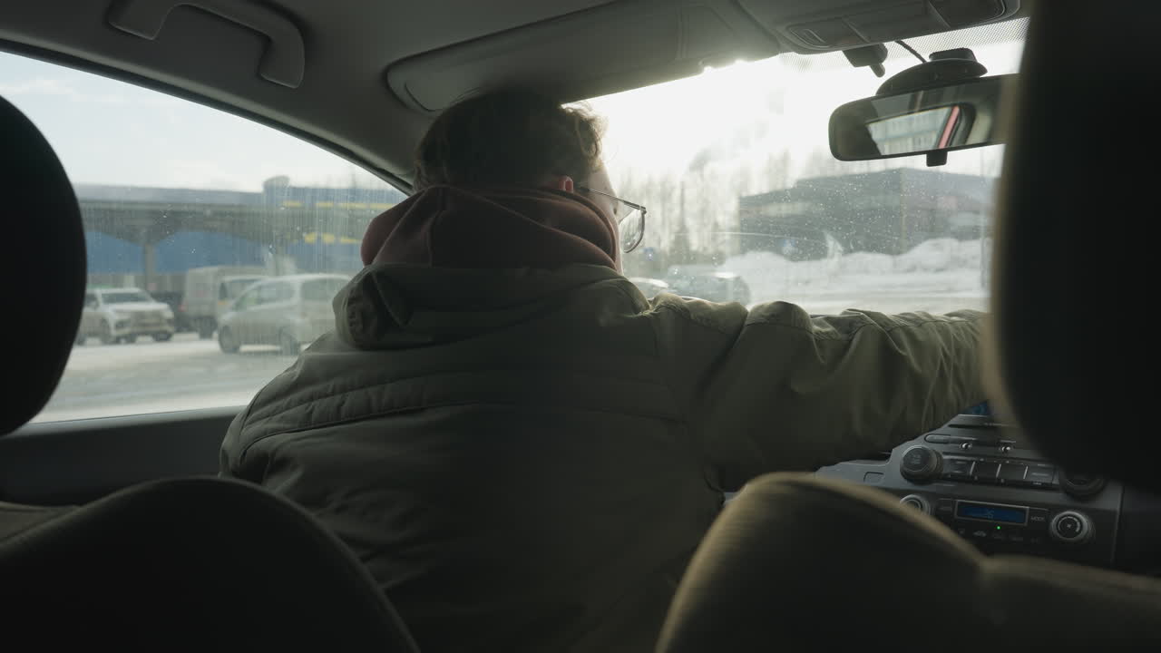 young man inside vehicle leans over passenger side while wiping car dashboard with blue cloth, snowy city street visible through window showing parked cars and large commercial building in background