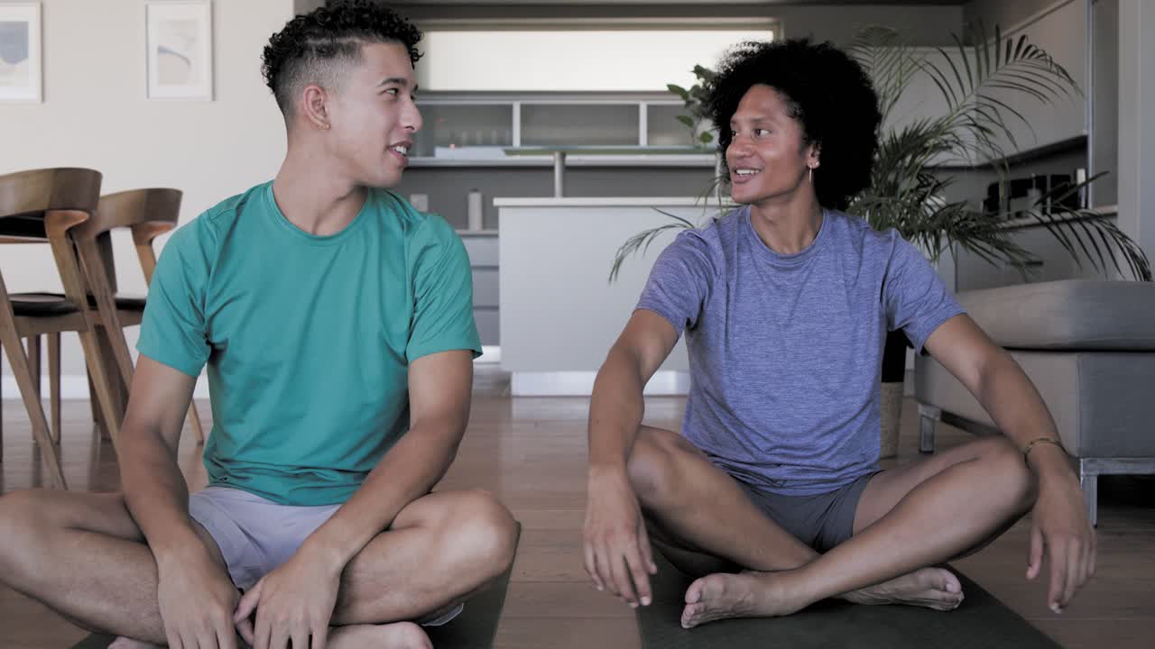 Male friends lowering hands after prayer, laughing on green mats with water bottles in living room