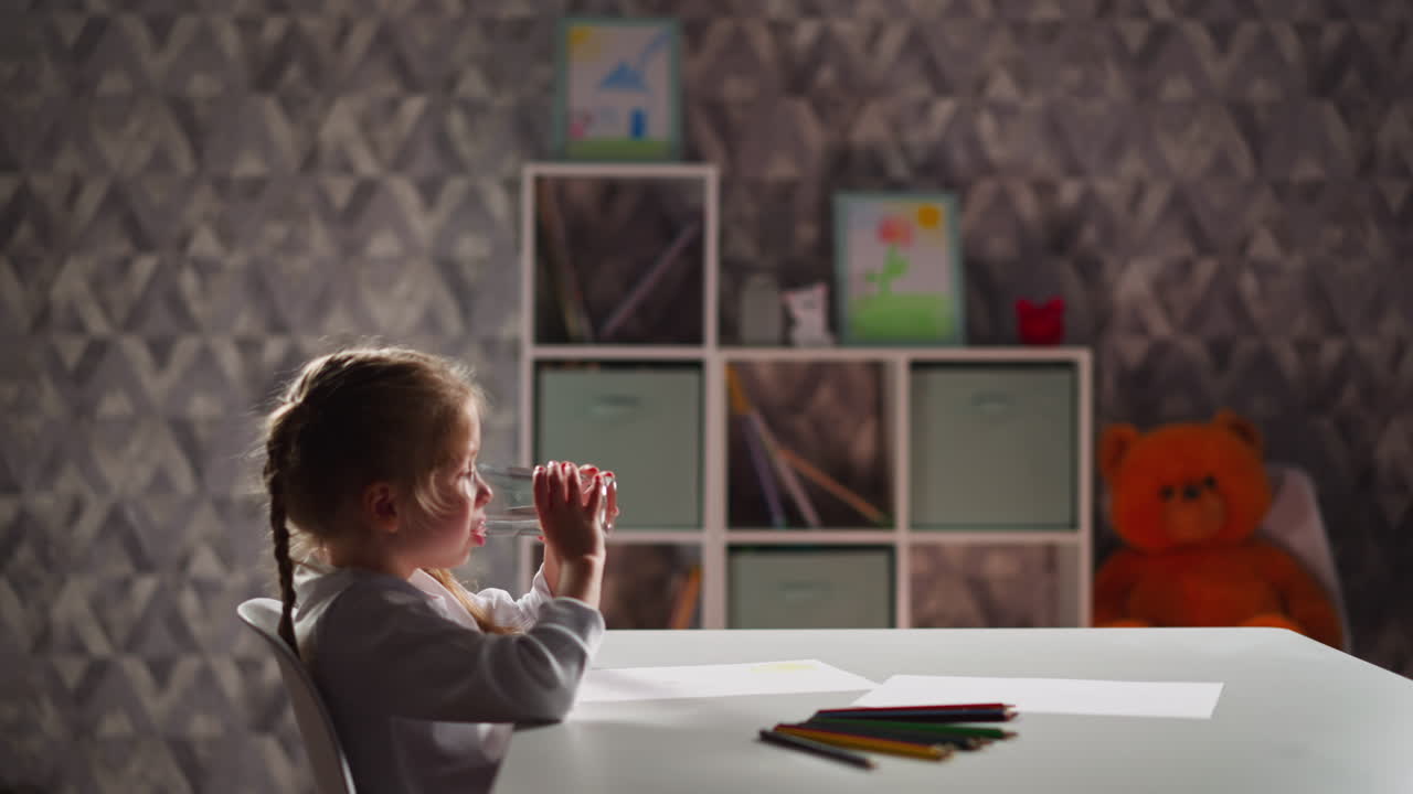 Una bambina carina beve acqua a tavola con carta e pastelli colorati.