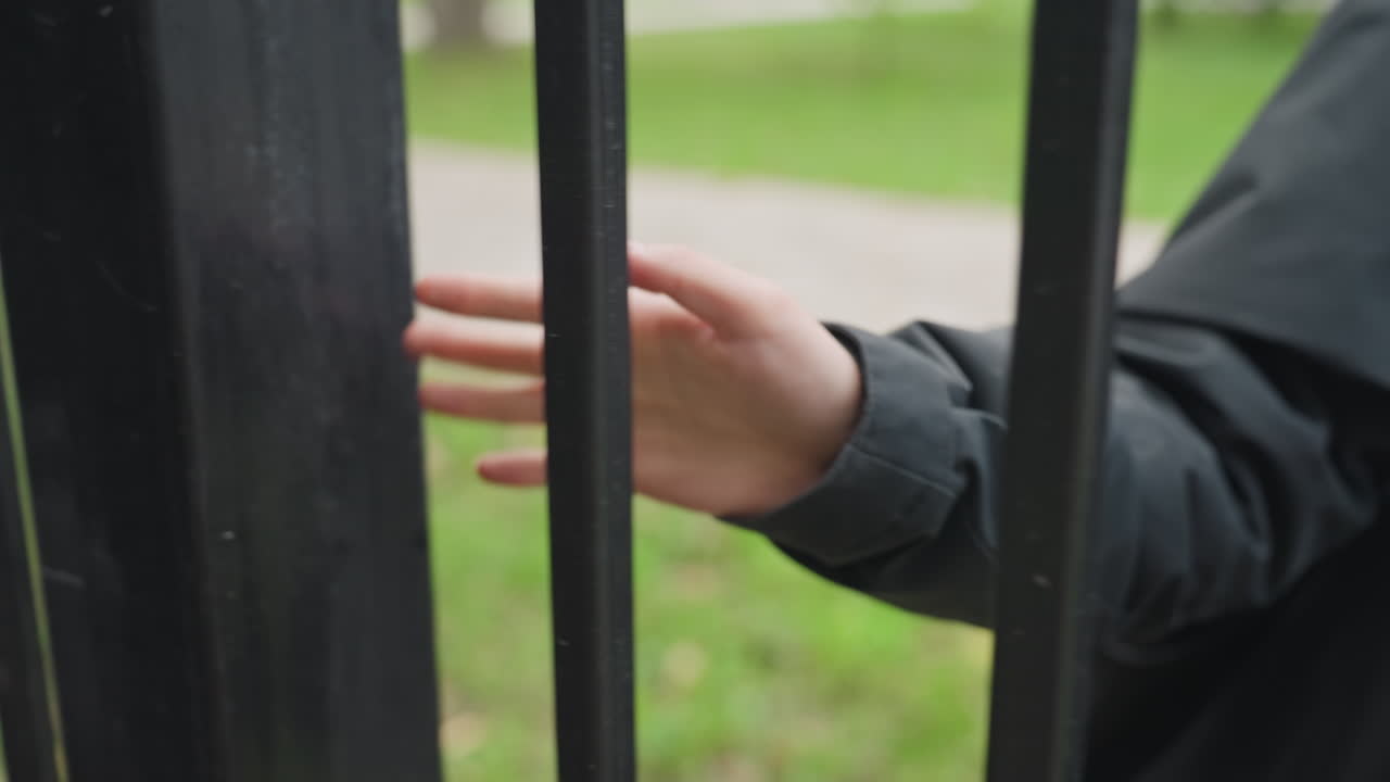 Mano che si allunga attraverso una recinzione di ferro, visitatore con giacca scura tocca decorazioni ornamentali in ferro battuto, sentiero del parco sfocato sullo sfondo, dita che scivolano lungo le sbarre con un gesto lento, evoca desiderio, restrizione