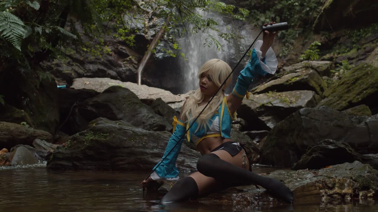 lucy heartfilia cosplayer sostiene su cuerda en posición mientras se sienta en la base de una cascada