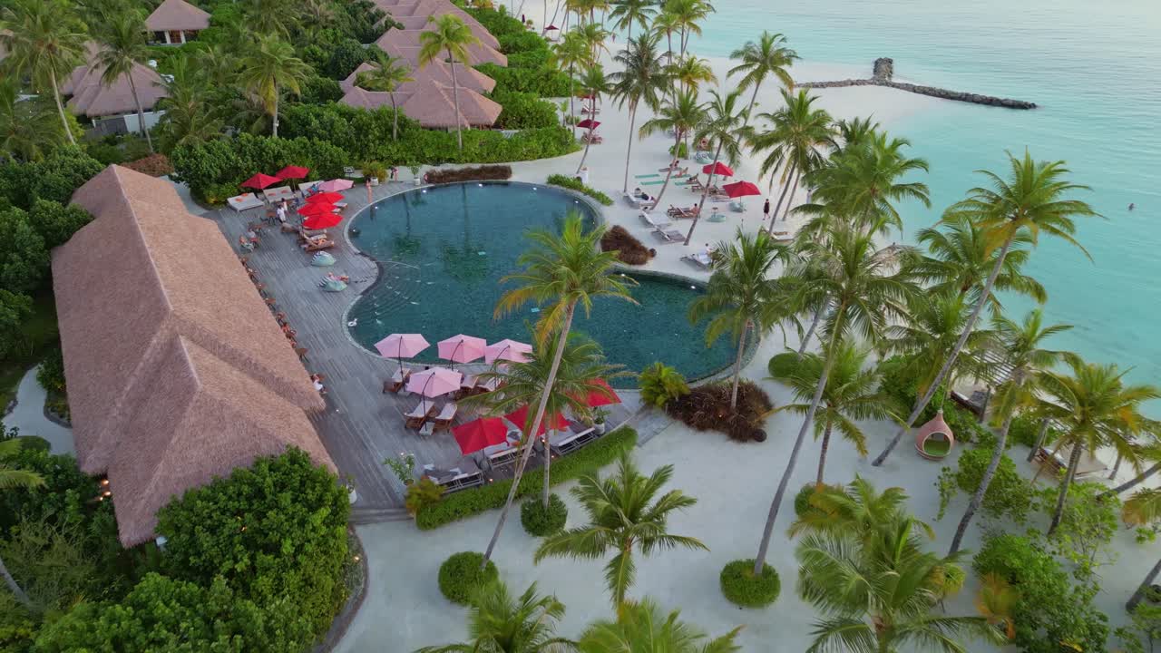 Swimming pool with palm trees and luxury resort hotel during sunny day. Aerial top down rising shot. Private sandy beach and ocean at sunset. Maldives, Asia.