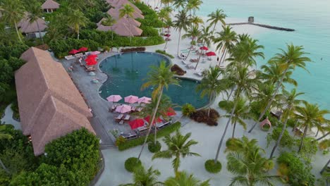 swimming pool with palm trees and luxury resort hotel during sunny day. aerial top down rising shot. private sandy beach and ocean at sunset. maldives, asia.
