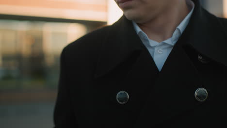 close-up of man holding phone to answer a call, wearing black suit with metallic buttons, white shirt underneath, set against blurred background with warm sunlight reflections on glass building