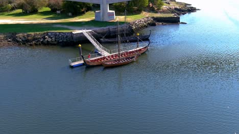 two people observe and browse on the jetty some viking ships restored imitation of the originals in the ulla river, sunny summer afternoon, drone shot orbits zenithal