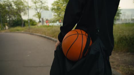 un primer plano de un hombre con una chaqueta negra y una mochila negra va a un juego de baloncesto con una pelota de baloncesto por la calle