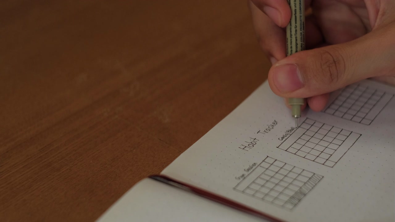 Habit Tracker Journal - Person's Hand Writing With A Pen On A Notebook With A Grid Pattern. - closeup shot