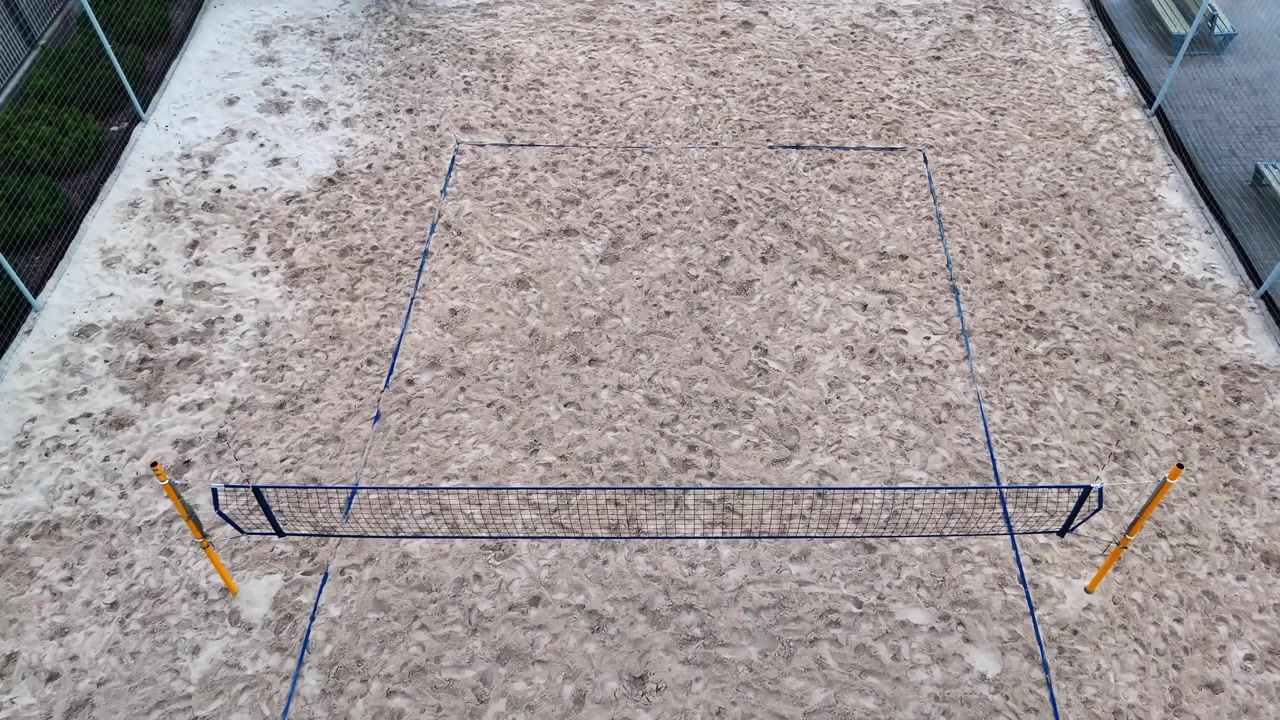Oblique drone view of an empty beach volleyball court with a taut net, blue boundary lines and footprint-textured sand inside a fenced urban sports facility.