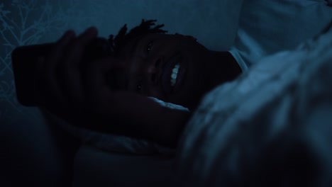 hombre sonríe mientras usa un teléfono inteligente en la cama por la noche