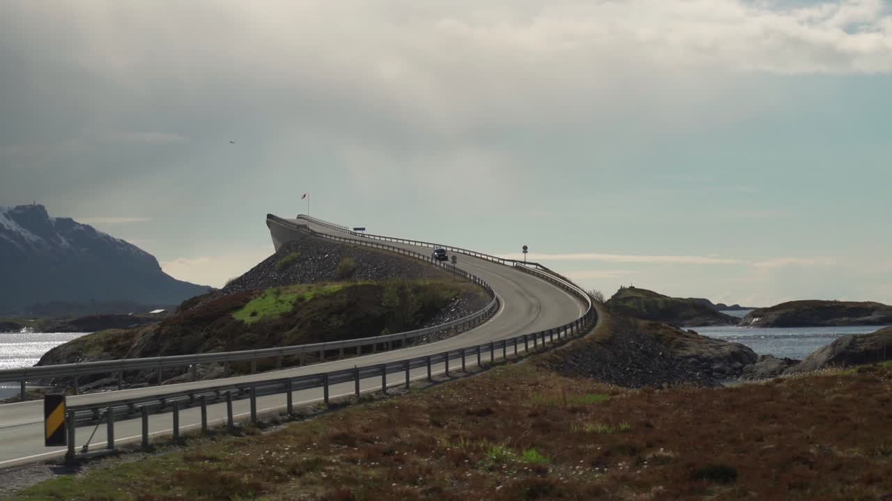 A view of the famous Atlantic Road in Norway. One of its'  curved bridges rises above the sea. It is overcast, gray haze hanging above the water. A dark silhouette of a mountain on the horizon