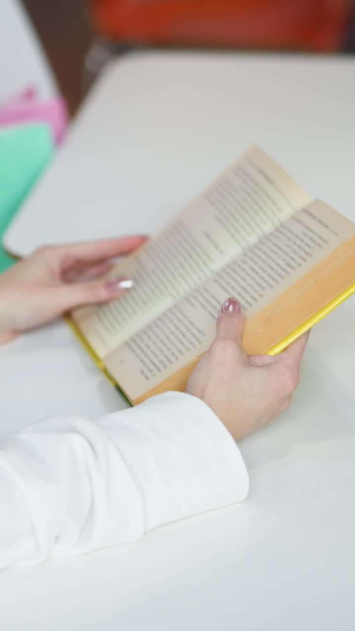 primer plano de una mujer sosteniendo un libro amarillo abierto mientras está sentada en una mesa en un centro comercial, una taza de café y bolsas de compras coloridas a su lado, con iluminación vibrante y fondo borroso