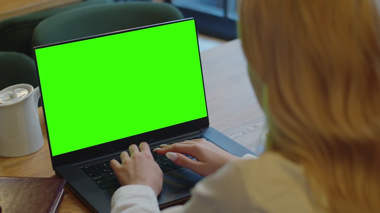 Over the shoulder shot of a business woman working in cafe interior on laptop on desk, looking at green screen. Woman using laptop computer with laptop green screen, sitting at wooden table.
