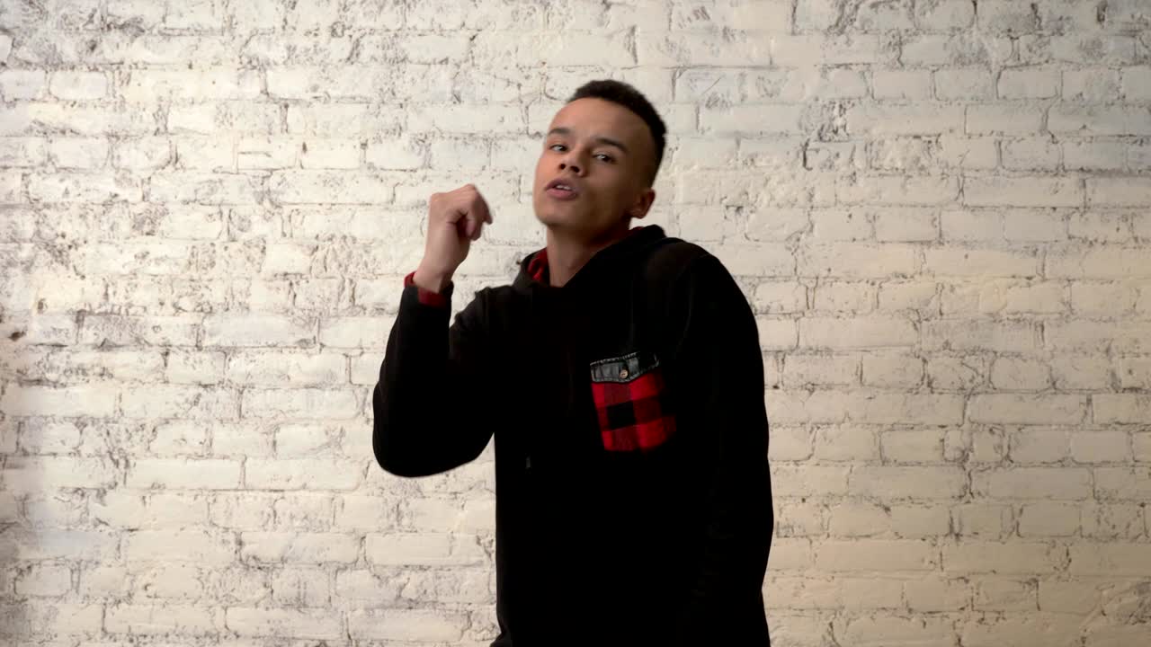 joven africano con una sudadera negra está bailando impetuosamente contra una pared blanca. discoteca, danza, movimiento, energía, concepto juvenil. bloquear en la cámara. 60 fps