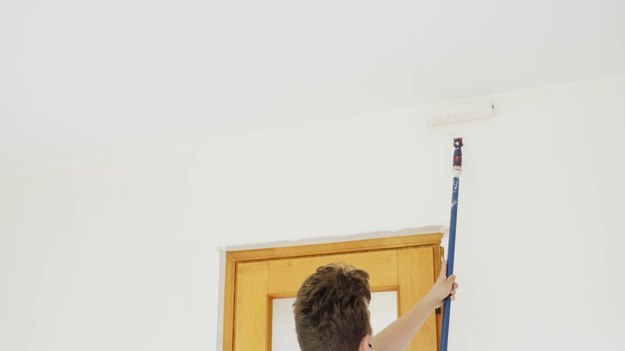 Person carefully applies white paint with a roller brush on an interior wall above a wooden door frame showcasing precise home renovation work in a residential living space