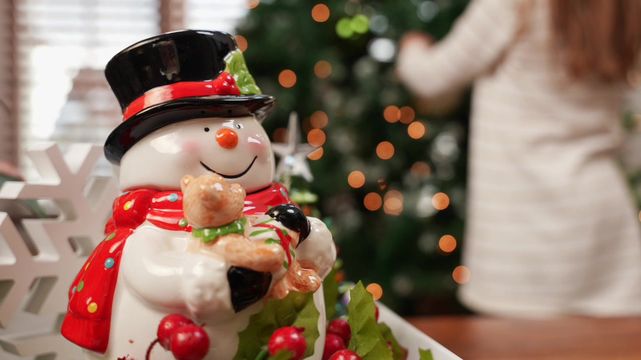 A medium shot of a glass snowman cookie jar in focus, with a 5-year-old girl hanging an ornament on a Christmas tree blurred in the background. A charming festive scene in 4K.