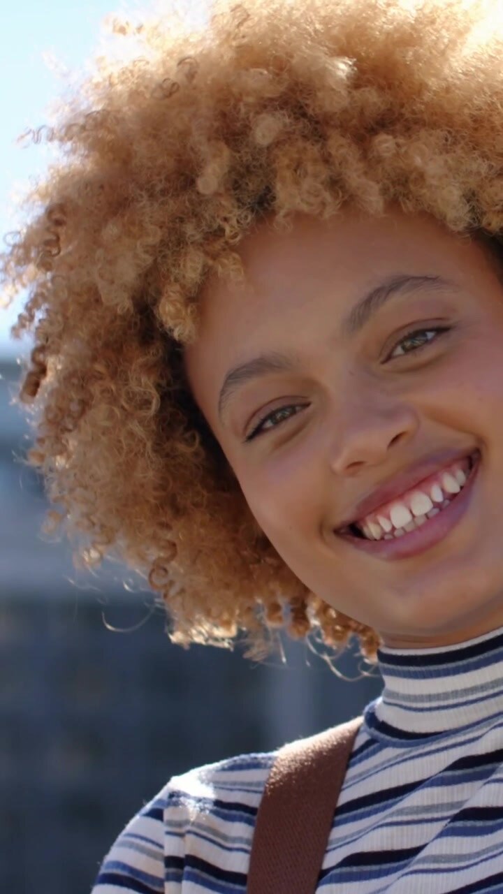Video verticale: Donna sorridente alla telecamera che si inclina a destra uscendo dalla strada, spallina del top a righe, spazio per il testo.
