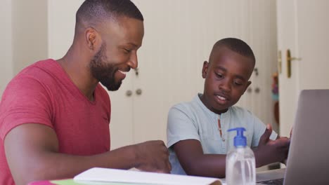 African-american-father-helping-son-with-homework-while-sitting-with-laptop-and-sanitizer-bottle-on-