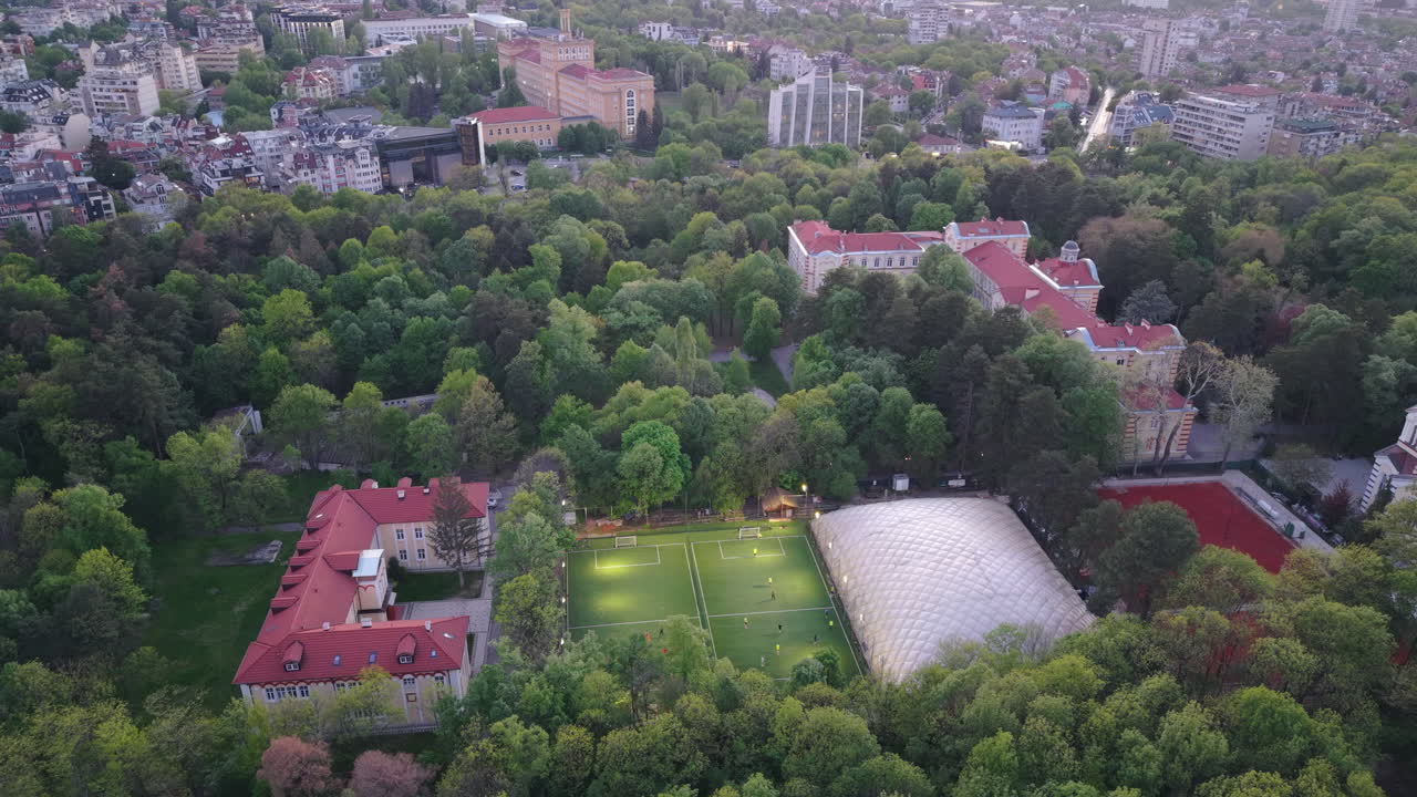 High-angle drone footage capturing a friendly soccer game in a public park in Sofia, Bulgaria. The top-down view shows players actively running on a green pitch, framed by lush spring trees.