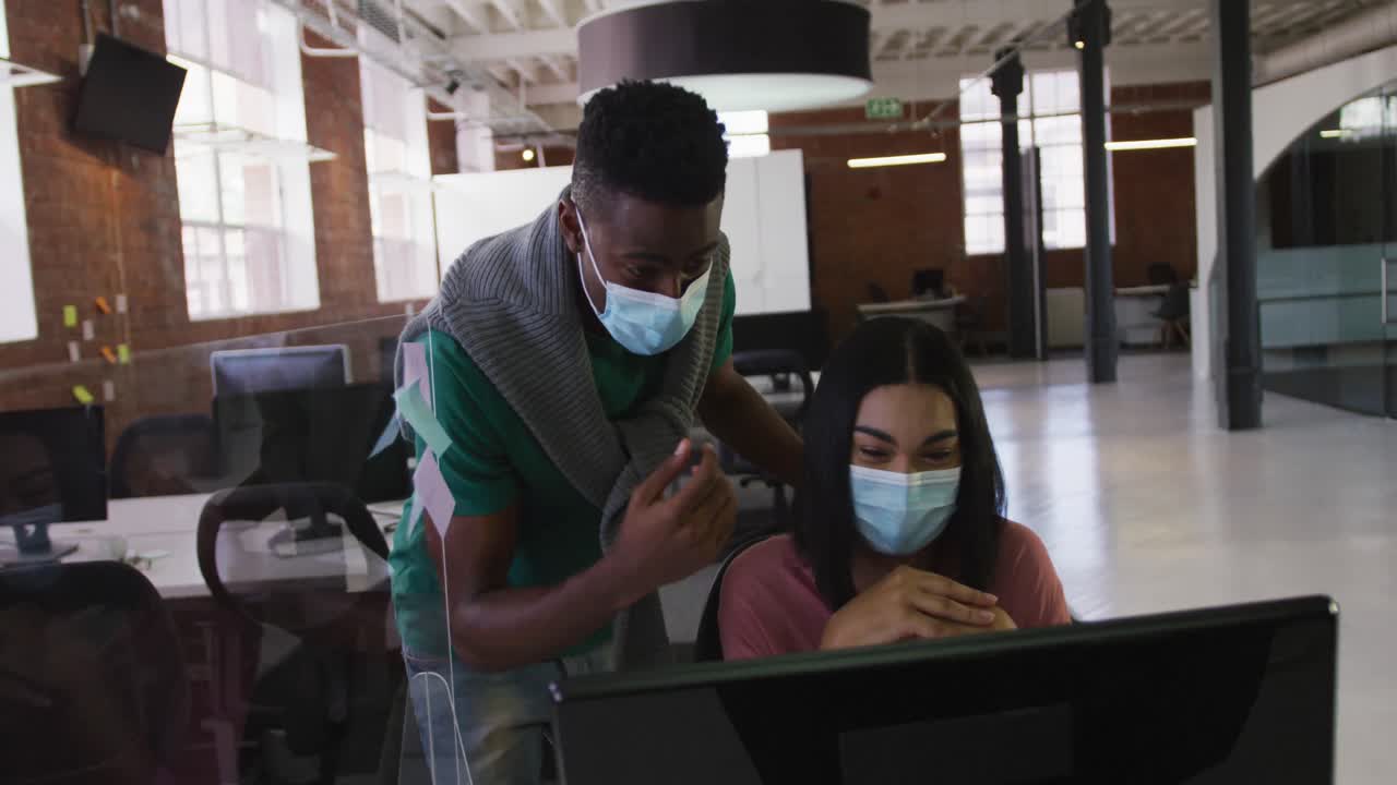 Diverse male and female business colleagues wearing face masks in discussion at work using computer