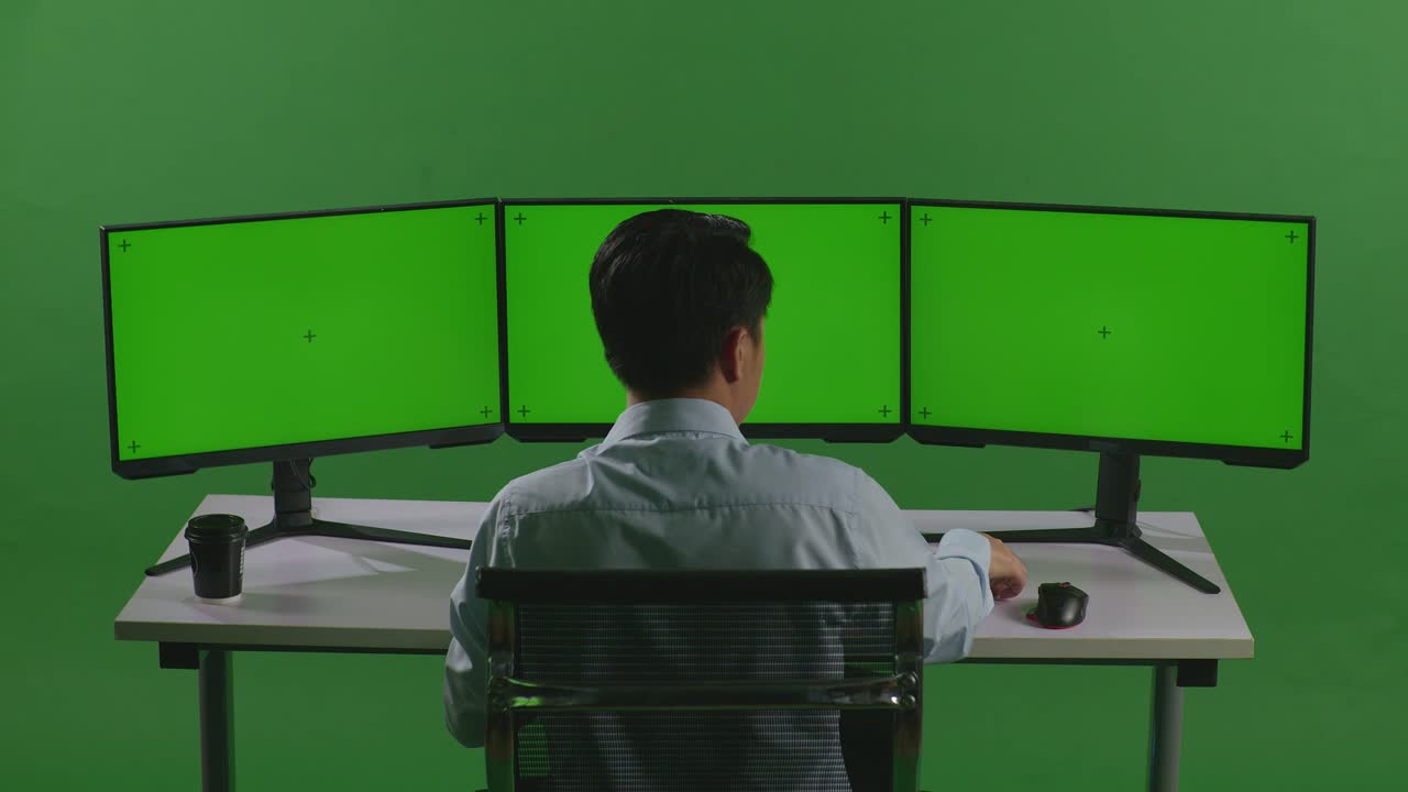 녹색 스크린 스튜디오에서 모의 여러 컴퓨터 모니터와 함께 작업하는 동안 휴대 전화로 이야기하는 아시아 남자의 뒷면