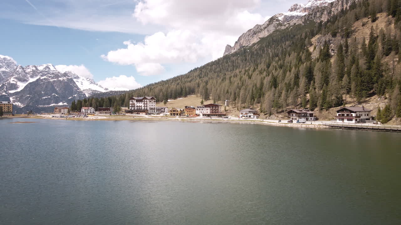 Camera flies steadily forward across a calm mountain lake, approaching a cluster of alpine lodges nestled at the forested shoreline beneath rugged, snow-dusted peaks