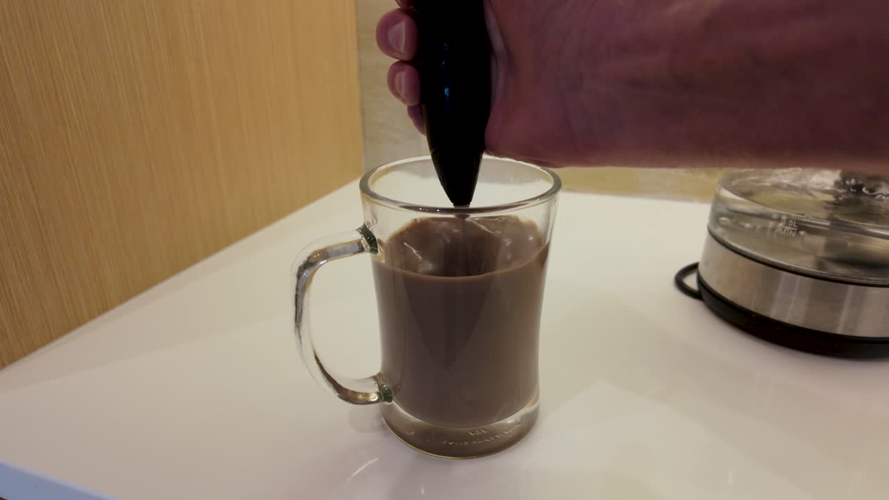 A close-up shot capturing the frothy swirl of instant hot chocolate in a clear mug as an electric whisk blends ingredients on a sleek countertop. Warm lighting enhances the cozy ambiance
