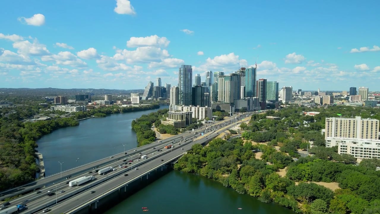 Imagens de drone da Interestadual 35 cortando o Centro de Austin, Texas, ao longo do Rio Colorado em um dia ensolarado. Apresenta tráfego, o horizonte da cidade e espaços verdes exuberantes.