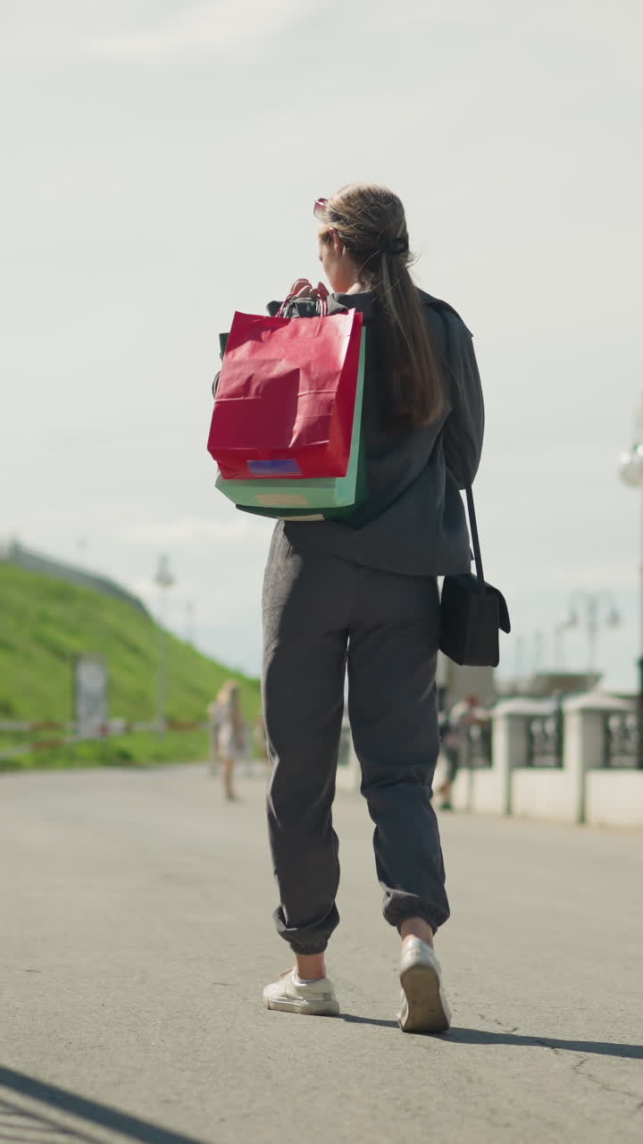 la señora disminuye la velocidad, ajustando las bolsas de compras en su hombro mientras camina cerca de las escaleras del puente, el fondo presenta una vista borrosa de la gente en la distancia, una colina y un paisaje pintoresco