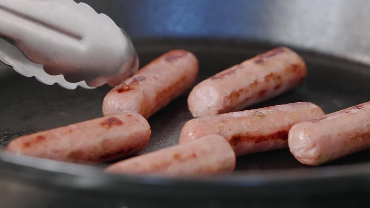 Tongs flipping breakfast sausages in hot cast iron pan to cook evenly