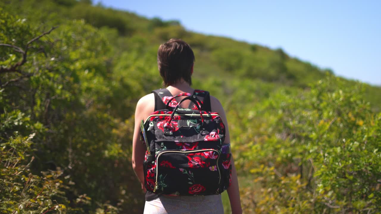 mujer niña dama con cabello corto y camiseta sin mangas, caminando o caminando por un exuberante parque público verde afuera con una mochila cubierta de rosas en un día caluroso y soleado durante el verano de 2021