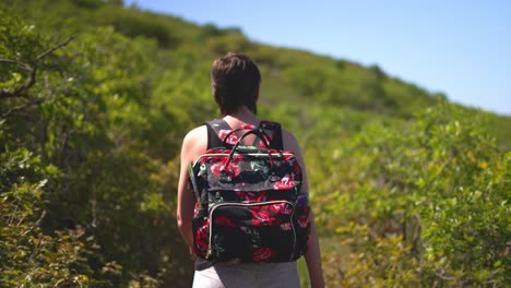 women girl lady with short hair and tank top on, walking or hiking lush green public park trail outside with rose covered backpack on a bright sunny hot day during summer in 2021