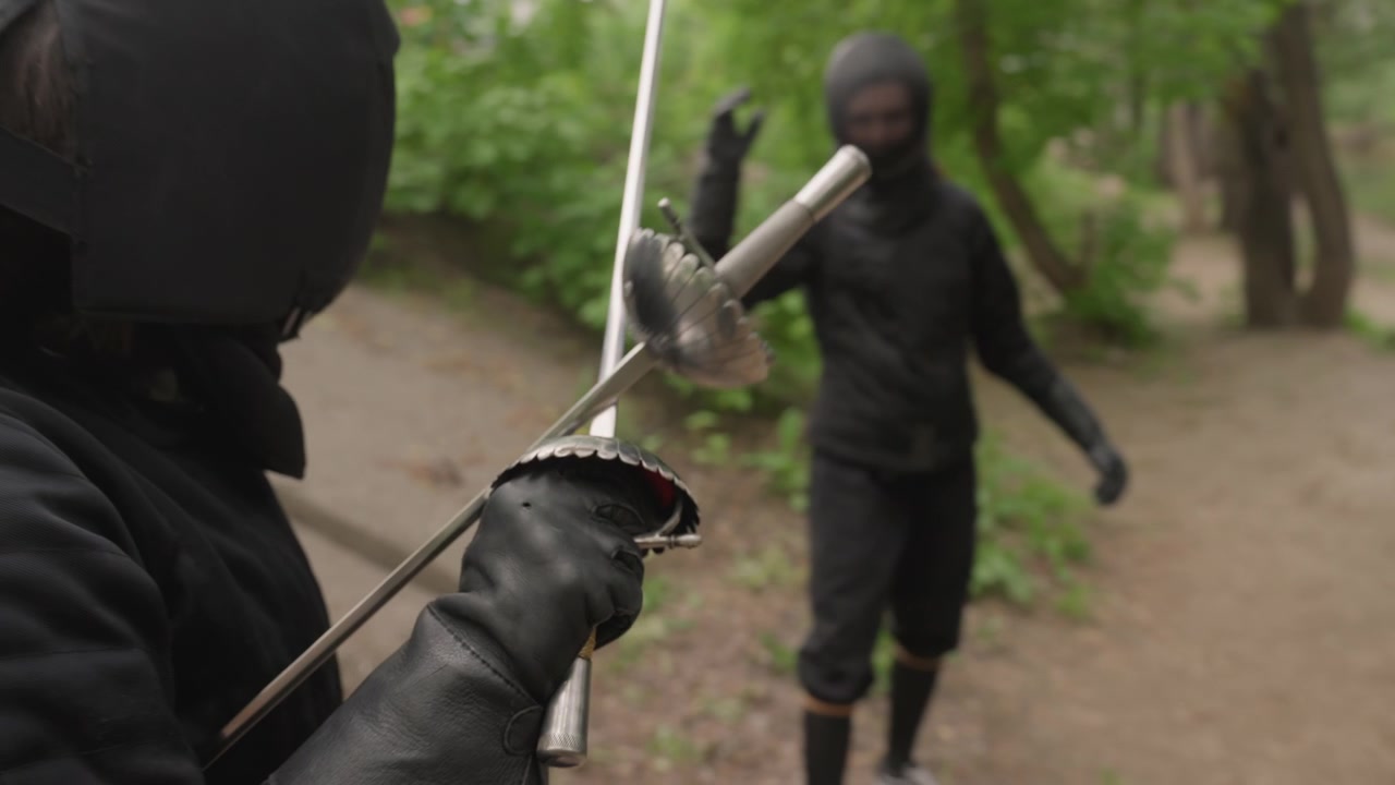 Close up of two people in black ninja attire practicing sword fighting outdoors on dirt path surrounded by greenery. One person holds camera while engaging with another both wearing