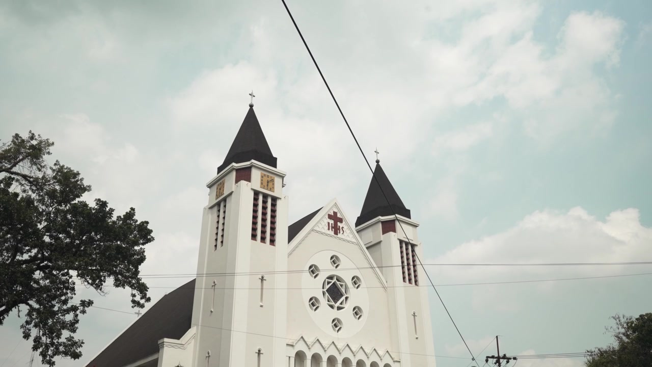 Vista ampla de catedral branca com torres gémeas e bandeiras numa rua tranquila em Malang.
